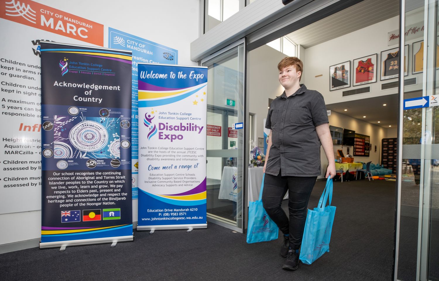 A young person carrying two blue promotional bags walks through the sliding glass entrance doors of an indoor venue, past large event banners.